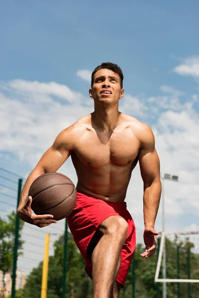 Jugador de baloncesto de carrera mixta sin camisa enfocado con pelota en la cancha de baloncesto bajo el cielo azul - foto de stock