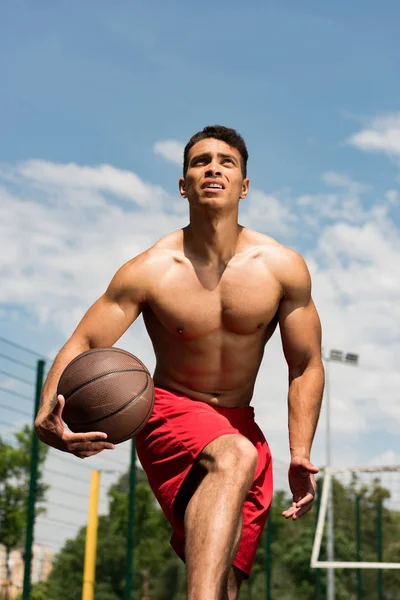 Foco sem camisa jogador de basquete de corrida mista com bola na quadra de basquete sob céu azul — Fotografia de Stock