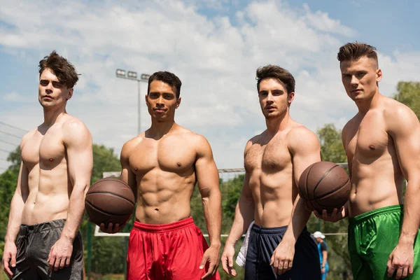 Cuatro jugadores de baloncesto sexy sin camisa con pelotas en la cancha de baloncesto en un día soleado - foto de stock