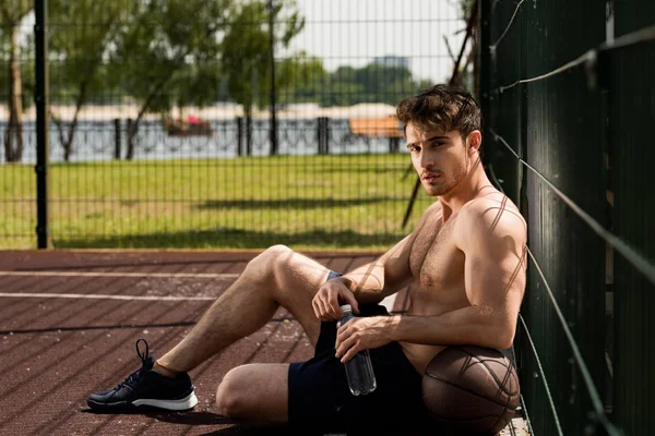 Sexy desportista muscular com bola e garrafa de água sentado no campo de basquete — Fotografia de Stock