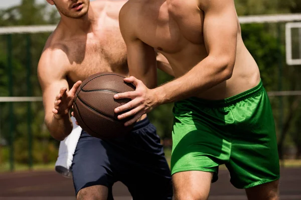 Vista parcial de dos deportistas sin camisa jugando baloncesto en cancha de baloncesto en un día soleado - foto de stock