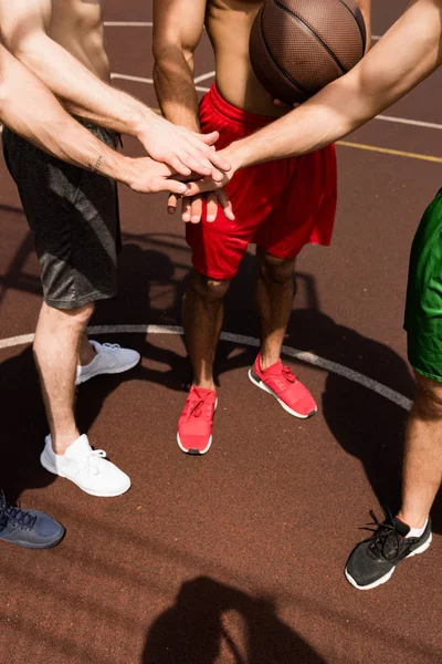 Vista recortada de cuatro jugadores de baloncesto sin camisa tocando las manos en la cancha de baloncesto en el día soleado - foto de stock
