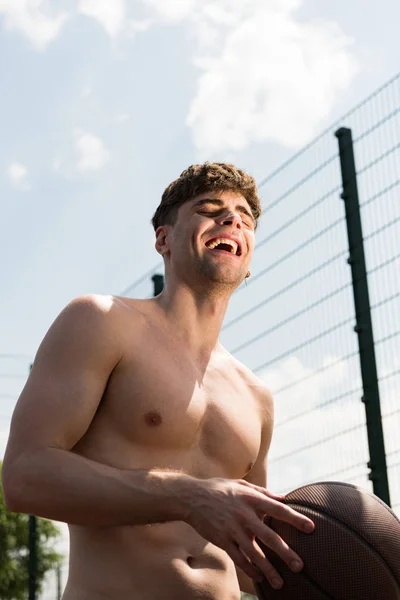 Sonriente sexy sin camisa deportista celebración de pelota en la cancha de baloncesto bajo el cielo azul - foto de stock