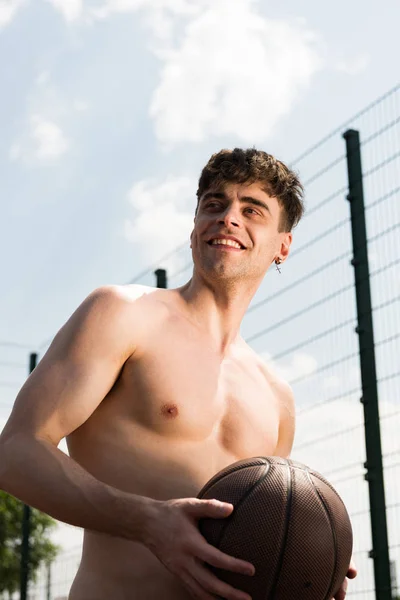 Усміхнений сексуальний спортсмен без сорочки тримає м'яч на баскетбольному майданчику під блакитним небом — стокове фото