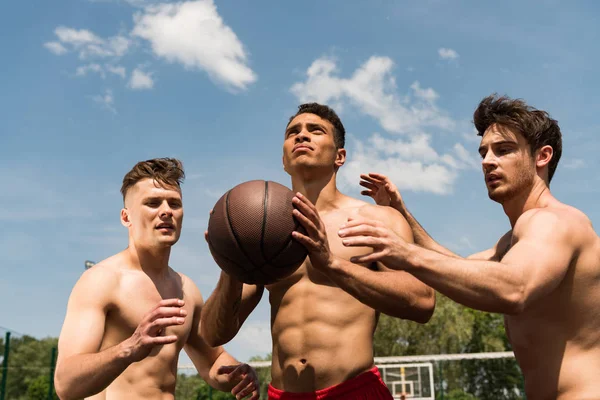 Tres deportistas sexy sin camisa jugando baloncesto bajo el cielo azul - foto de stock