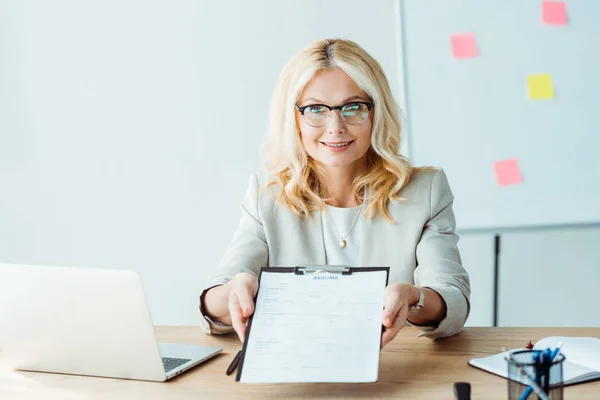 Blonde fröhliche Frau mit Brille hält Klemmbrett mit Lebenslauf, während sie am Schreibtisch sitzt — Stockfoto