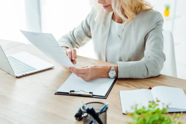 Обрезанный вид женщины, смотрящей на бумагу возле ноутбука и буфера обмена в офисе — стоковое фото