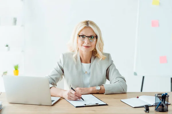 Cheerful woman holding pen near clipboard and laptop in office — Stock Photo