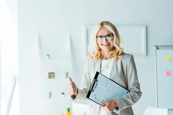 Fröhliche blonde Frau mit Brille, die Daumen nach oben zeigt, während sie Klemmbrett hält — Stockfoto