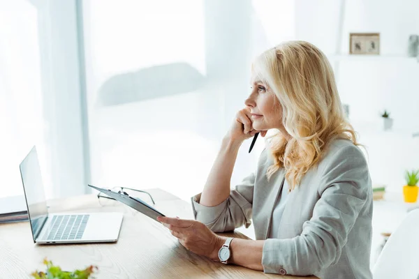 Thoughtful blonde woman holding clipboard and pen near laptop in office — Stock Photo