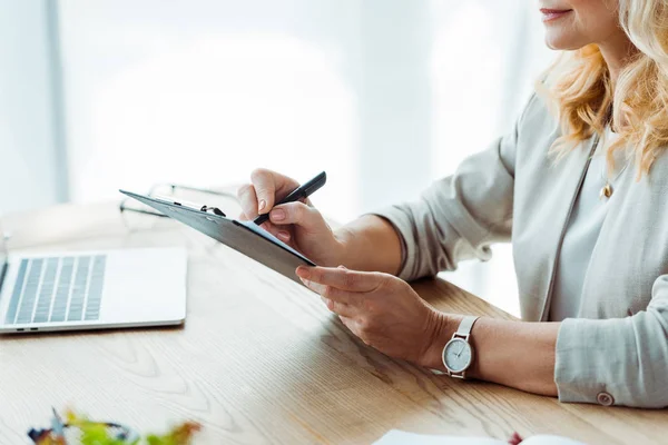 Cropped view of woman writing while holding clipboard in office — Stock Photo