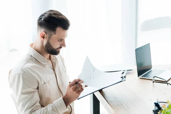 Hombre barbudo guapo mirando el papel mientras sostiene el portapapeles - foto de stock