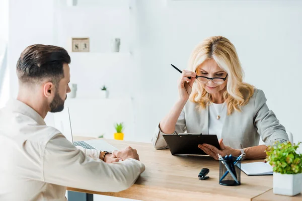Selective focus of blonde recruiter in glasses looking at clipboard near bearded man — Stock Photo