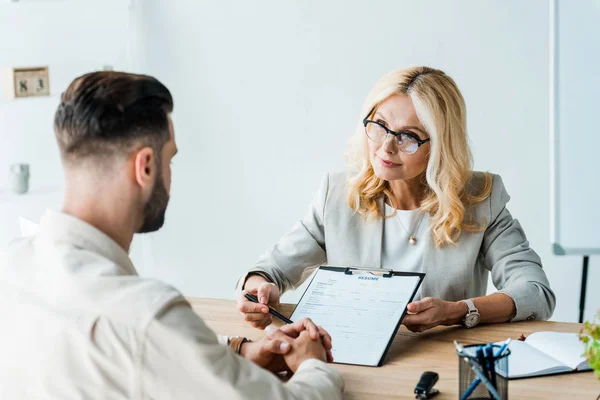 Enfoque selectivo del reclutador en gafas que sostienen la pluma cerca del portapapeles y mirando al hombre barbudo - foto de stock