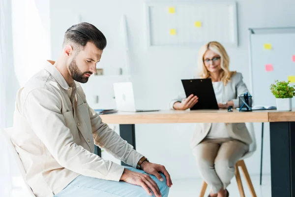 Selective focus of upset man sitting near blonde woman with clipboard — Stock Photo