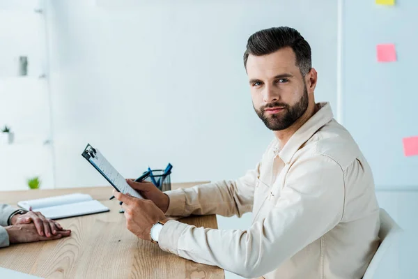 Abgeschnittene Ansicht eines Personalvermittlers in der Nähe eines gutaussehenden bärtigen Mannes, der in die Kamera schaut, während er Klemmbrett hält — Stockfoto