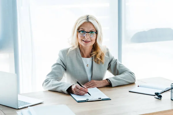 Fröhliche blonde Frau mit Brille blickt auf Klemmbrett neben Laptop — Stockfoto