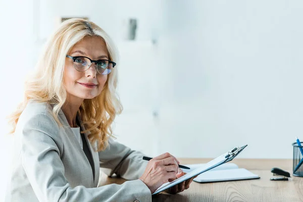 Atractiva mujer rubia en gafas sosteniendo pluma y portapapeles - foto de stock