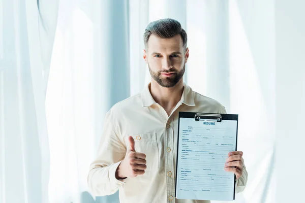 Guapo barbudo mostrando el pulgar hacia arriba y sujetando el portapapeles con letras de curriculum vitae - foto de stock