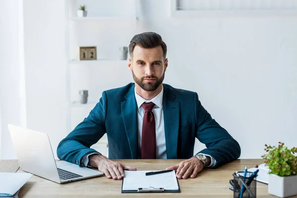 Schöner Personalvermittler sitzt in der Nähe von Klemmbrett und Laptop im Büro — Stockfoto