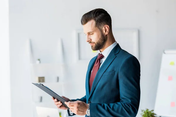 Bel homme barbu en costume regardant le presse-papiers dans le bureau — Photo de stock