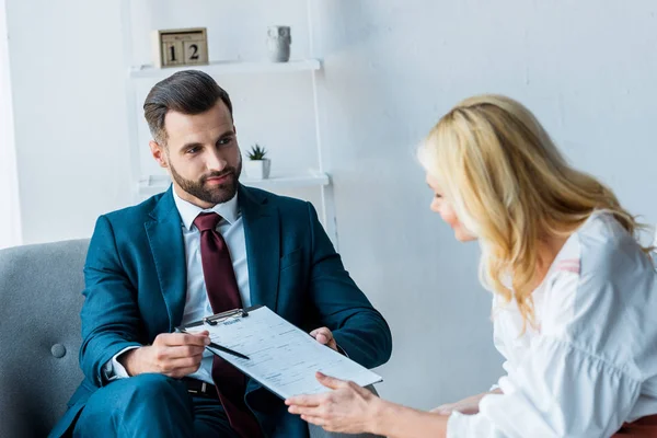 Selective focus of handsome recruiter holding clipboard and pen while sitting in armchair near blonde employee — Stock Photo