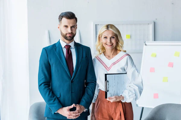 Reclutador guapo con las manos apretadas y mujer rubia con portapapeles de pie en la oficina - foto de stock