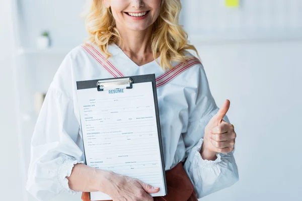 Vista recortada de la mujer rubia feliz sujetando portapapeles con letras de curriculum vitae y mostrando el pulgar hacia arriba - foto de stock