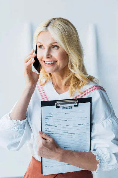 Mujer rubia feliz hablando en el teléfono inteligente y sujetando portapapeles con letras de curriculum vitae - foto de stock
