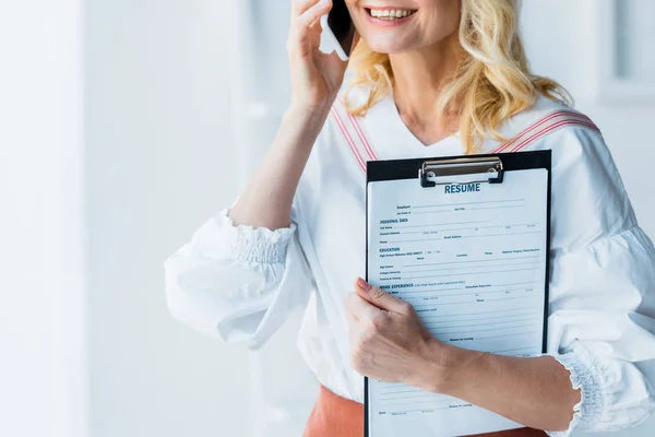 Vista recortada de c mujer rubia alegre hablando en el teléfono inteligente y sujetando portapapeles con letras de curriculum vitae - foto de stock