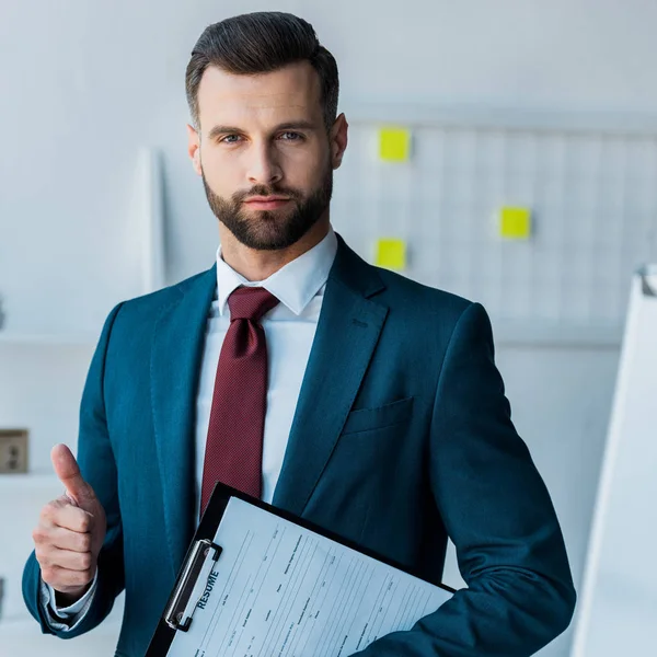 Hombre barbudo confiado sosteniendo portapapeles con letras de curriculum vitae y mostrando el pulgar hacia arriba - foto de stock