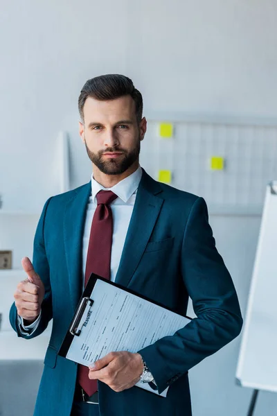 Serious bearded man holding clipboard with resume letters and showing thumb up — Stock Photo