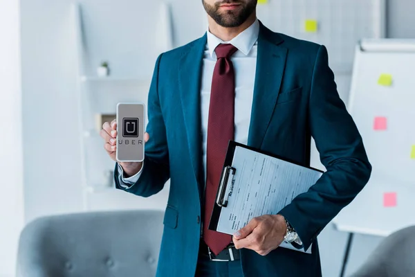 KYIV, UCRANIA - 10 de junio de 2019: vista recortada del hombre barbudo sujetando el portapapeles y el teléfono inteligente con la aplicación uber en la pantalla - foto de stock