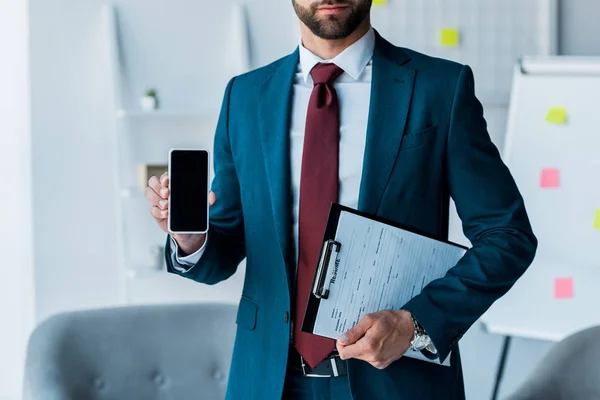 Vista recortada del hombre barbudo sosteniendo teléfono inteligente con pantalla en blanco y portapapeles - foto de stock