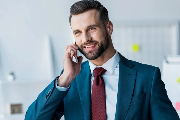 Fröhlicher bärtiger Mann im Anzug, der im Büro mit dem Smartphone spricht — Stockfoto