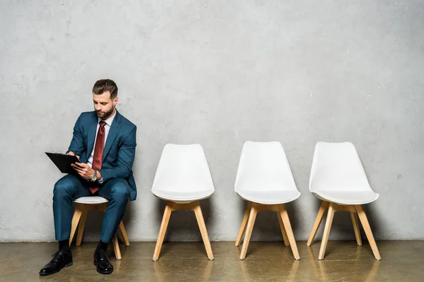 Schöner Mann sitzt auf weißem Stuhl und hält Klemmbrett — Stockfoto