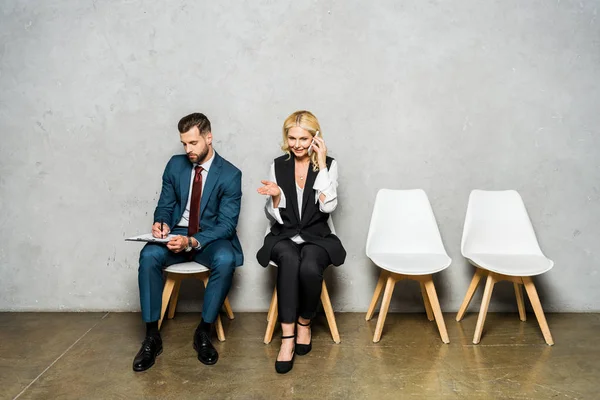 Glückliche blonde Frau, die auf dem Smartphone spricht, während sie neben Mann sitzt, der schreibt und Klemmbrett hält — Stockfoto