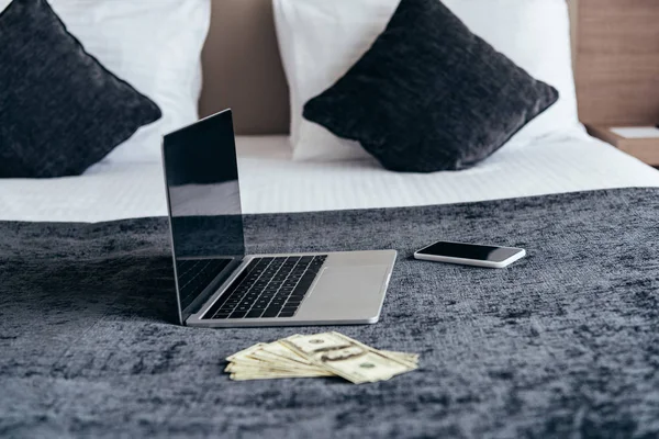 Laptop mit leerem Bildschirm, Smartphone und Dollarnoten auf dem Bett im Hotelzimmer — Stockfoto