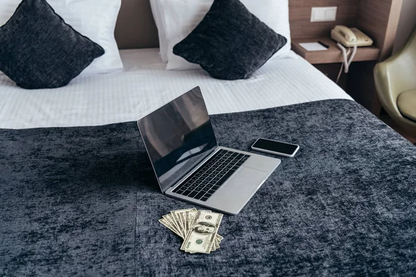 Ноутбук с чистым экраном, смартфоном и долларовыми банкнотами на кровати в номере отеля — стоковое фото