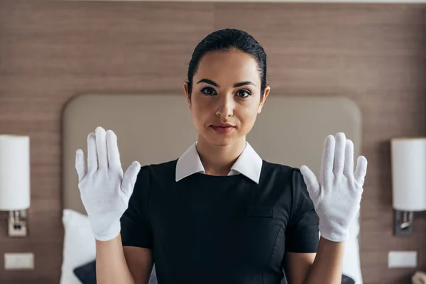 Vorderansicht des hübschen lächelnden Dienstmädchens in weißen Handschuhen und Schürze, das in die Kamera schaut und die Hände im Hotelzimmer hochhält — Stockfoto