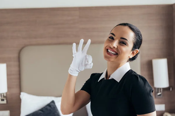 Souriant jolie femme de ménage en gant blanc montrant signe correct et regardant la caméra dans la chambre d'hôtel — Photo de stock