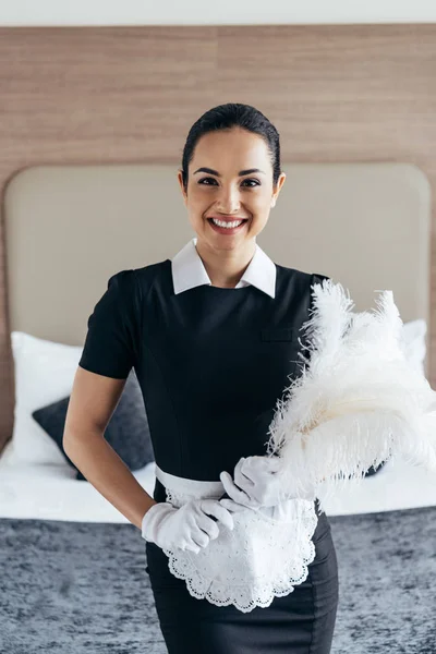 Vista frontal de empregada sorridente em luvas brancas segurando espanador perto da cama no quarto do hotel — Fotografia de Stock