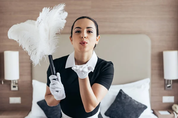 Vista frontal de la criada sonriente en guantes blancos sosteniendo el plumero y enviando un beso de aire cerca de la cama en la habitación del hotel - foto de stock