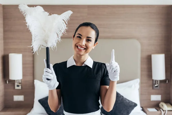 Vista frontal de la criada sonriente con guantes blancos sosteniendo el plumero y mostrando el cartel de la idea cerca de la cama en la habitación del hotel - foto de stock