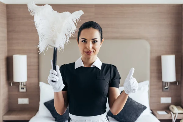 Vue de face de la bonne souriante en gants blancs tenant le plumeau et montrant pouce vers le haut près du lit dans la chambre d'hôtel — Photo de stock