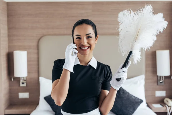 Vista frontal de la sonrisa bonita sirvienta en guantes blancos sosteniendo el plumero y hablando en el teléfono inteligente en la habitación de hotel - foto de stock