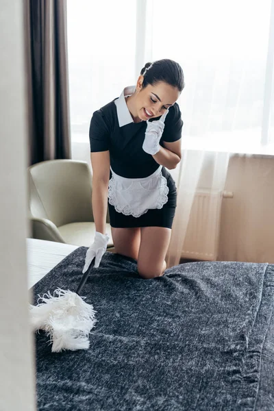 Lächelnd hübsches Dienstmädchen in weißen Handschuhen, das Bett mit Staubwedel säubert und im Hotelzimmer mit dem Smartphone spricht — Stockfoto