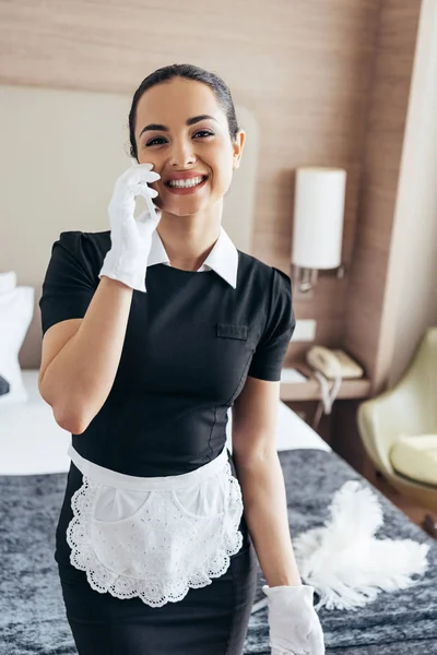 Sonriente criada en guantes blancos y delantal hablando en smartphone en habitación de hotel - foto de stock