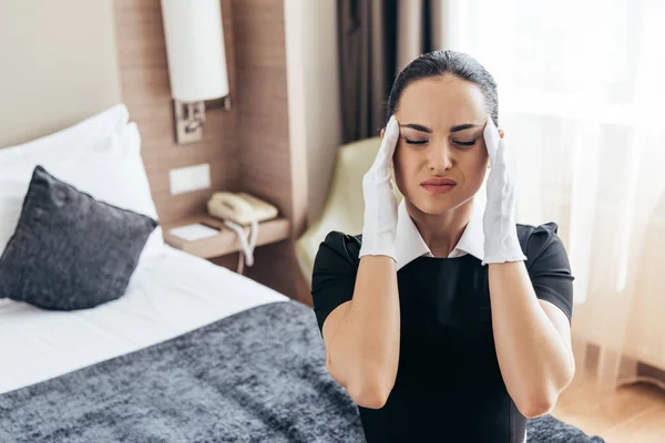 Müdes Dienstmädchen in weißen Handschuhen sitzt mit geschlossenen Augen auf dem Bett und berührt den Kopf — Stockfoto