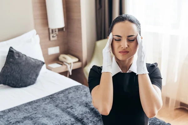 Cansada criada en guantes blancos sentada en la cama con los ojos cerrados y tocando la cabeza - foto de stock